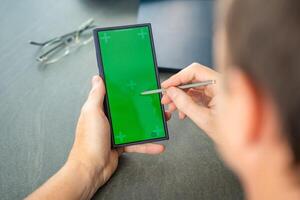 cerca arriba ver de hombre mano participación inteligente teléfono con verde croma llave pantalla y aguja bolígrafo a su creativo espacio de trabajo foto