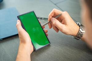 Close up view of man hand holding smart phone with green chroma key screen and stylus pen at his creative workspace photo