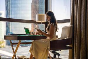 Stylish businesswoman freelancer working with laptop and smartphone, she is sitting on chair, enjoying panoramic view in the city background. Low key photo. High quality photo