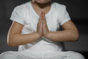 Tres años antiguo pequeño niña meditando en un loto actitud en un gris antecedentes en oscuro habitación. alto calidad foto