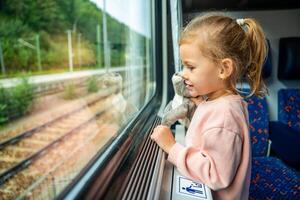 pequeño niña con juguete mirando fuera tren ventana afuera, mientras eso Moviente. de viaje por ferrocarril en Europa foto