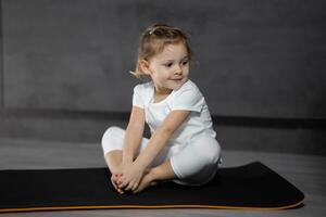 pequeño linda niña practicando yoga actitud en gris antecedentes en oscuro habitación. alto calidad foto