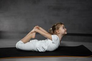 pequeño linda niña practicando yoga actitud en gris antecedentes en oscuro habitación. alto calidad foto