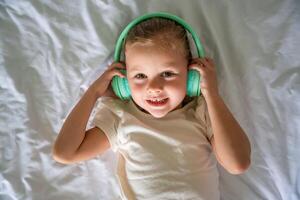 Cute little girl listening music using green kids headphones headphones in home bed. High quality photo