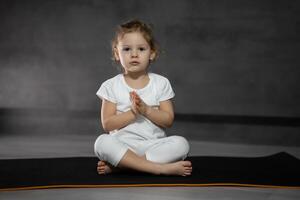 Three years old little girl meditating in a lotus pose on a gray background in dark room. High quality photo