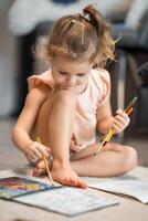 Little girl sits on the floor at home and draws with paints and brushes in a coloring book. Early childhood creativity and education. High quality photo