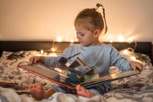 Little girl reading pop up book in home bed in Christmas environment with lights background. High quality photo