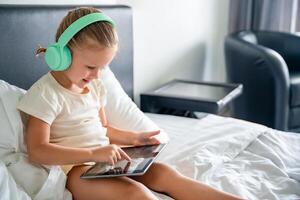 linda pequeño niña en auriculares utilizando digital tableta y sonriente contento mientras escuchando a música o jugando juego en hogar cama. alto calidad foto