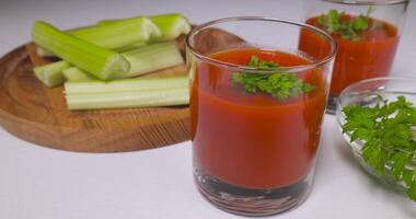 Organic freshly squeezed tomato juice into a glass, celery and parsley on a white table. Healthy and vegetarian food concept. Ultra 4K video