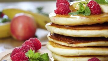 Closeup of Pouring honey on stack of pancakes. Tasty breakfast food. Pancakes are served with raspberries, banana and mint leaf. Ultra 4K video