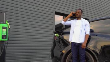 nero uomo parlando su il Telefono mentre ricarica lusso elettrico auto a stazione video