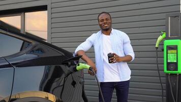 African-American man drinking coffee near electric car, waiting for it to charge at a charging station video