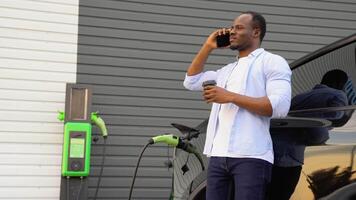 Black man talking on the phone and drinking coffee while charging luxury electric car at station video