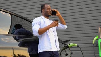 Happy young black man charging automobile battery from small public station, standing near electric car, drinking coffee and talking on smartphone video