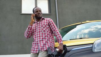 svart man talande på de telefon medan laddning frakt bil på elektrisk fordon laddning station video