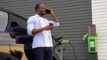 Black man talking on the phone and drinking coffee while charging luxury electric car at station video