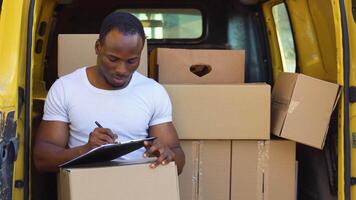 African american good looking deliveryman at the yellow van with carton boxes filling in documents on the clipboard video