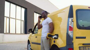 africano americano hombre hablando en el teléfono mientras cargando carga coche a eléctrico vehículo cargando estación video