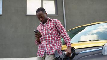 negro hombre participación teléfono inteligente mientras cargando camión coche a eléctrico vehículo cargando estación video