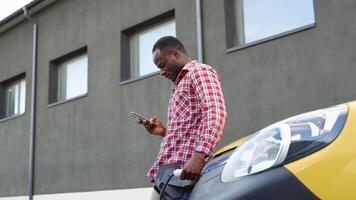 africano americano homem conversando em Móvel telefone enquanto elétrico carro é cobrando em ao ar livre estação video