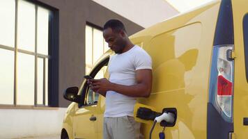 Black man chatting on mobile phone while electric car is charging on outdoor station video