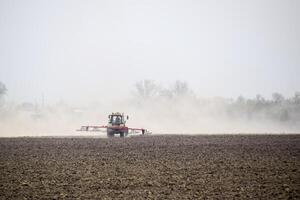 The tractor harrows the soil on the field and creates a cloud of dust behind it photo