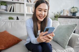 felizmente relajado joven mujer se sienta en un sofá utilizando un móvil teléfono, escuchando a música mediante un aplicación y auriculares foto