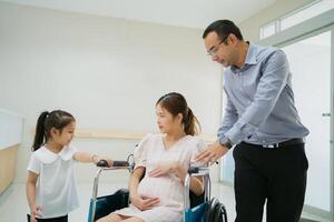 asiático marido tomando cuidado de embarazada mujer sentado en silla de ruedas y niños sonrisa a hospital foto