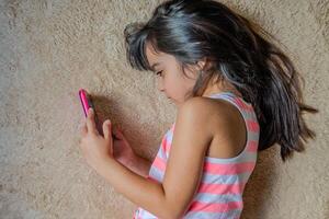 Cute little girl using a smartphone. Kid playing with mobile phone, lying on a bed. Freetime. Technology and internet concept. Toddler laying on bed and looking at smartphone. photo