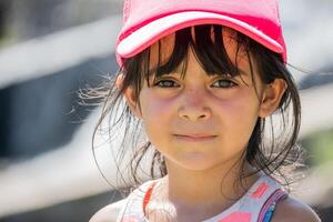 Very close portrait of a latin kid on a trip. The girl wears a pink cap. She looks at the camera with a very serious face. photo