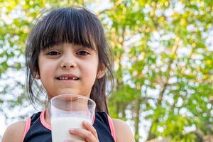 de cerca retrato de su. ella bebidas un vaso de frío Leche después jugando fuera de su casa. foto