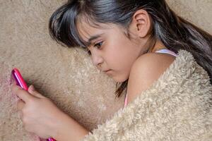 Cute little girl using a smartphone. Kid playing with mobile phone, lying on a bed. Freetime. Technology and internet concept. Toddler laying on bed and looking at smartphone. photo