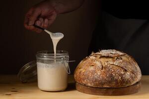 The hands of a young man showing what the sourdough is like in a glass jar. With a crusty sourdough bread next to him. photo