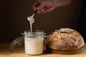 The hands of a young man showing what the sourdough is like in a glass jar. With a crusty sourdough bread next to him. photo