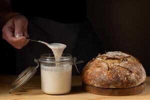 The hands of a young man showing what the sourdough is like in a glass jar. With a crusty sourdough bread next to him. photo