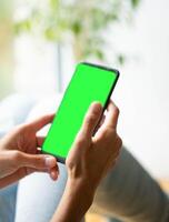 Blurred image of young woman sitting in relaxed pose. Smartphone with green screen in hands close up photo