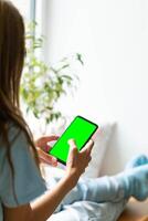 Young woman sitting on window skill at home with smartphone with green screen in hands. Soft focus. photo