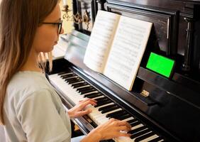 joven niña sentado a negro piano utilizando teléfono inteligente con verde pantalla a estudiando jugando piano. foto