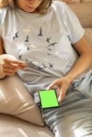 Young woman making online payment sitting at home with smartphone with green screen and bank card. photo