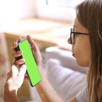Young woman in eyeglasses looking at green screen smartphone in hands sitting at home near window. photo