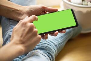 Close up smartphone with green screen in hands of sitting young girl in blue jeans, socks Soft focus photo