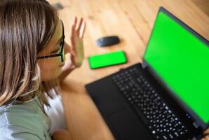 Young girl greets by raising hand, working, studying or connecting, looking at green screen laptop. photo