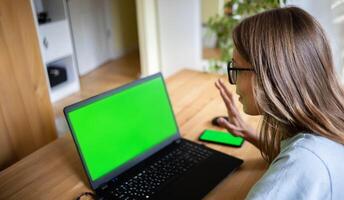 Young girl greets by raising hand looking at green screen laptop online working studying connecting photo