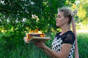 girl holds a burning book in her hands. A young woman in a forest burns a book. photo