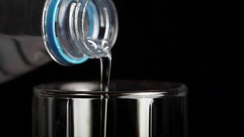 Water poured from the bottle into a transparent glass cup on a black background. Water is poured into a glass from bottle, close up video