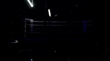 Boxing ring and two chairs with table dark background. View of a regular boxing ring surrounded by blue ropes spotlit by a spotlight. Light show around the boxing ring. video