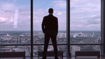 Full length back view of successful businessman in suit standing in office with hands on waist, CEO. Businessman from the back in front of a city view on the window video