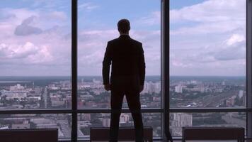 Businessman in suit standing next to a big window from an office building and checking his watch back view. Businessman standing at window looking on his wrist watch video