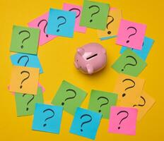 Ceramic piggy bank and stickers with drawn question marks on a yellow background, top view photo