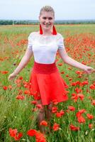 Blonde young woman in red skirt and white shirt, red earrings is in the middle of a poppy field. photo